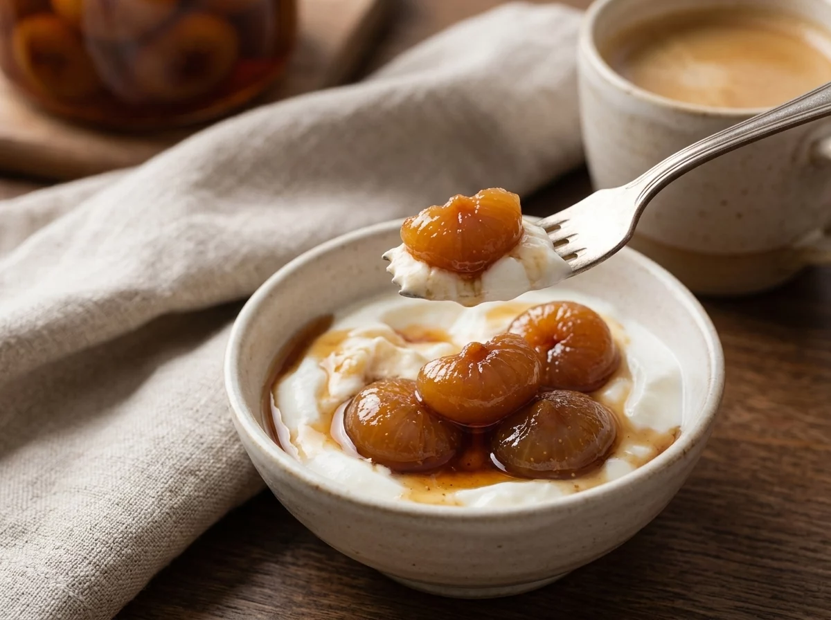 Bowl of yogurt topped with fig spoon sweet and syrup next to a coffee cup on a linen-covered table.